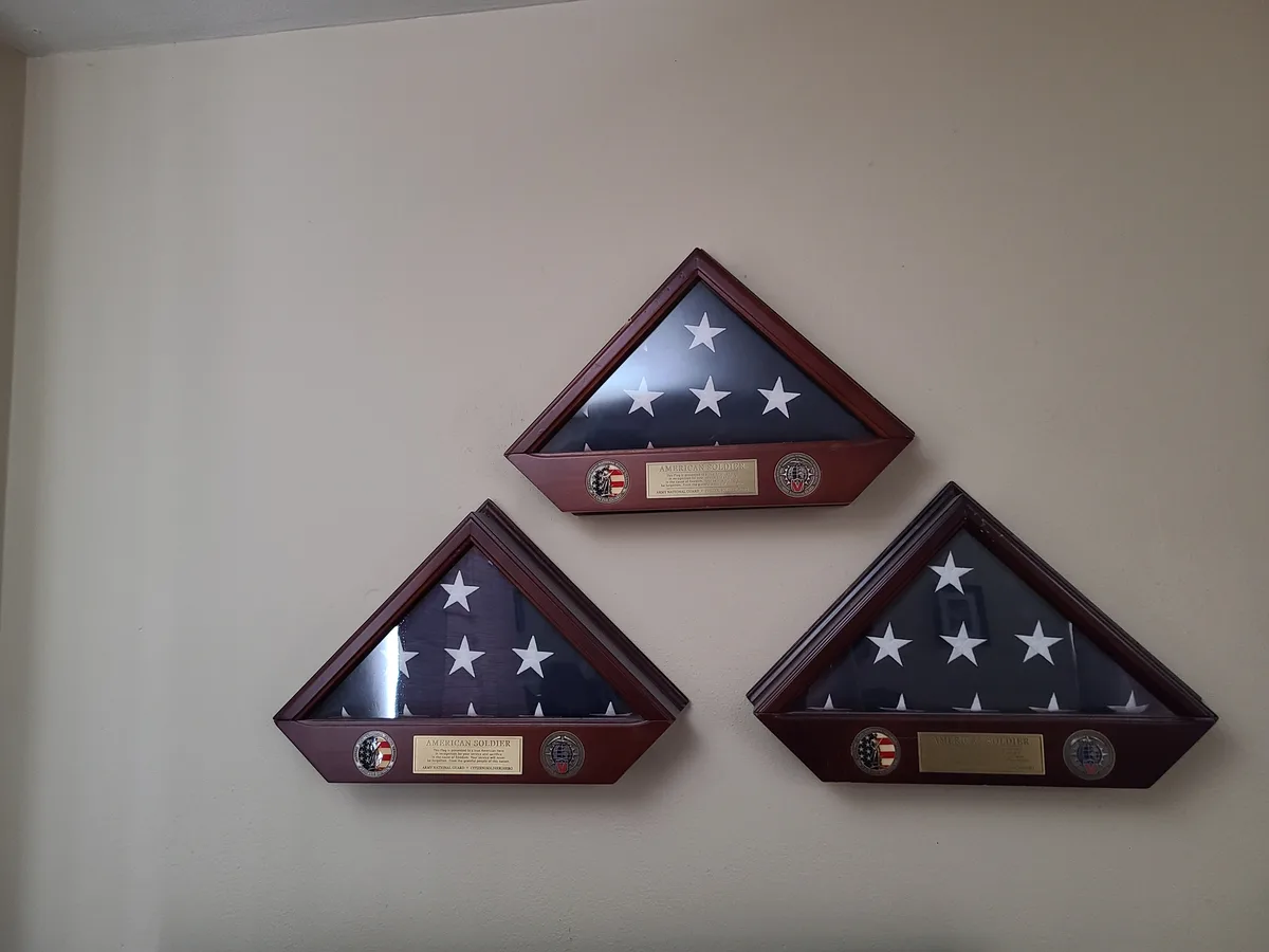 Three triangular American flags, folded and displayed in wooden frames on a beige wall, each with a plaque reading 'AMERICAN…