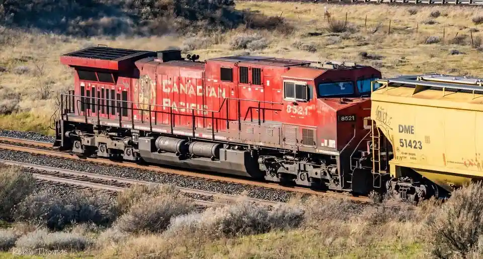 A red Canadian Pacific locomotive, numbered 8521, is shown on railway tracks in a dry, grassy landscape, pulling a yellow…