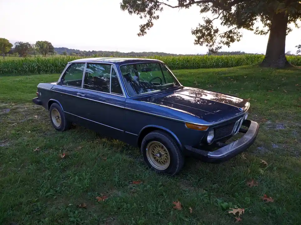 A dark blue classic BMW sedan with gold wire wheels is parked on grass under a tree, with a cornfield in the background. The…