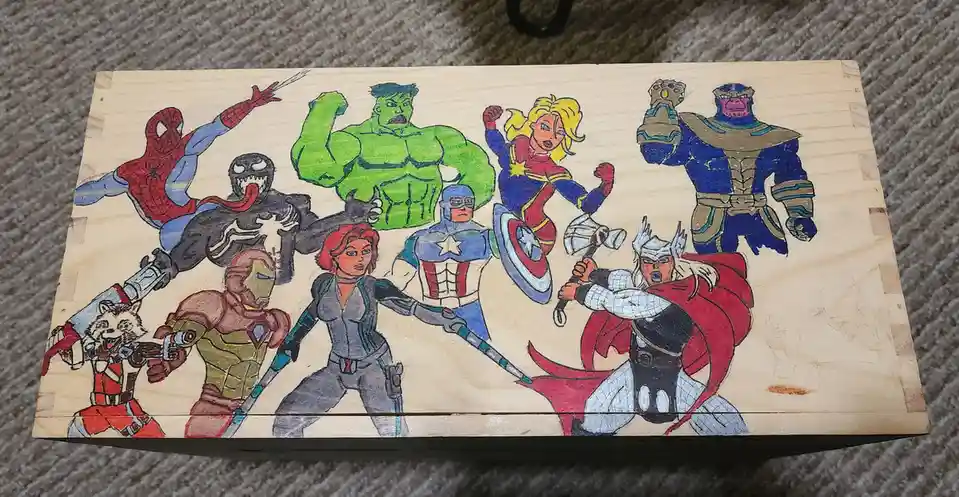 A hand-drawn illustration on a wooden box depicts a group of Marvel superheroes and villains, including Spider-Man, Venom…
