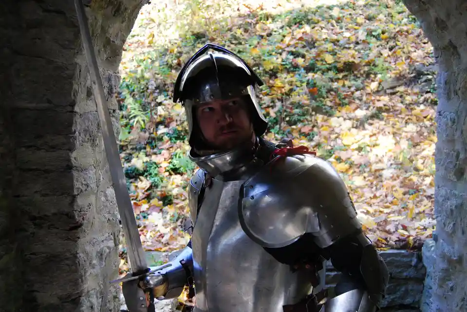 A man in full metallic medieval armor, including a helmet and breastplate, stands in a stone archway holding a sword. He…