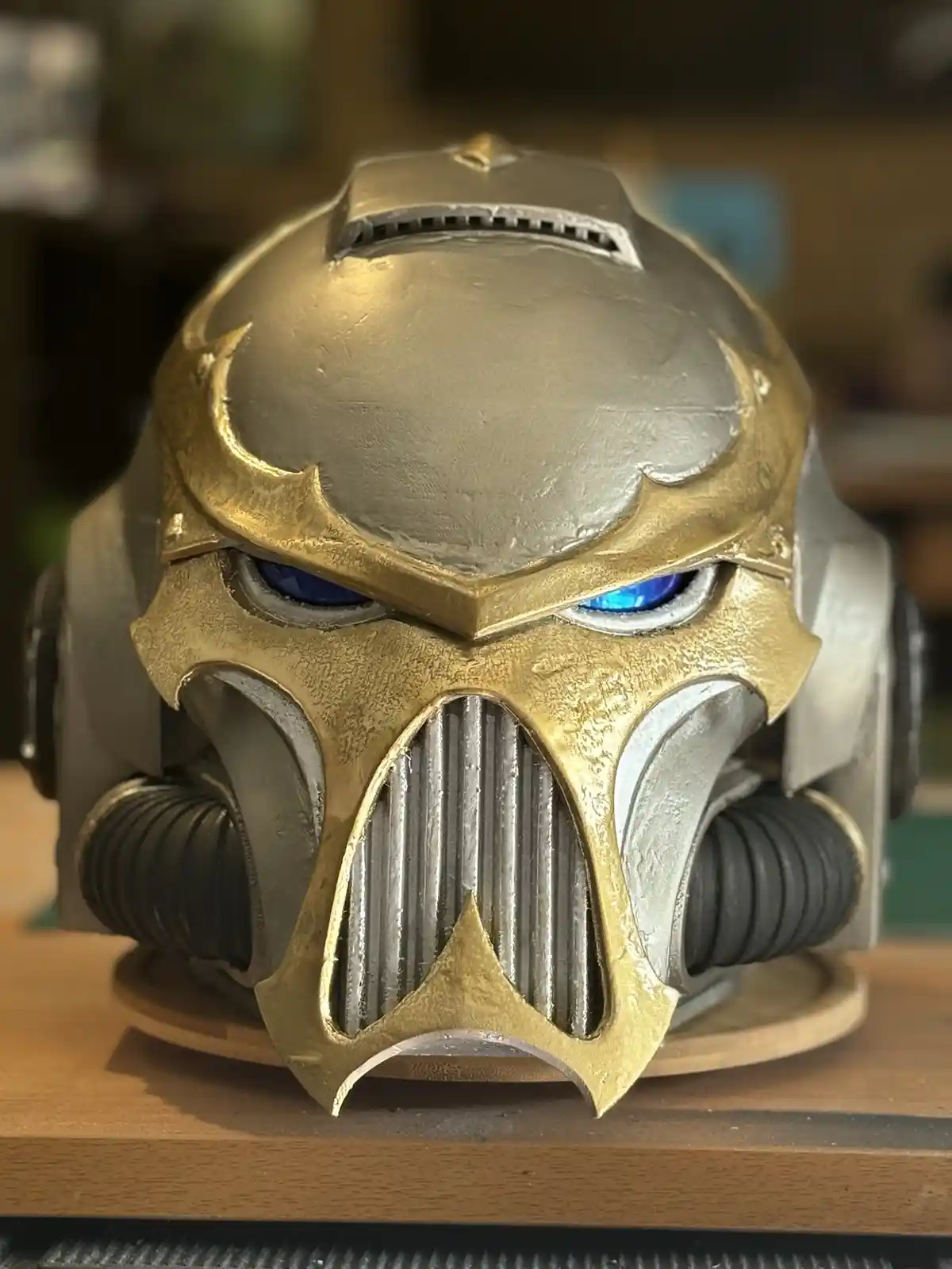 A close-up photo shows a detailed, handcrafted sci-fi helmet with gold and silver metallic paint, glowing blue eyes, and a…
