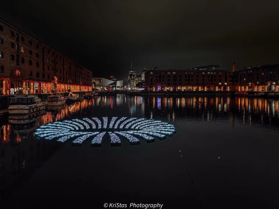 A nighttime photo of a waterfront with historic brick buildings and festive lights along the docks, reflecting in the dark…