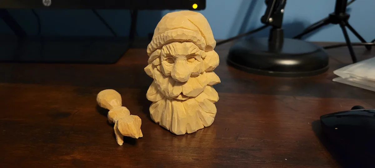 A rough wooden carving of a bearded man's head with a hat sits on a desk next to a small, unfinished wooden piece shaped…