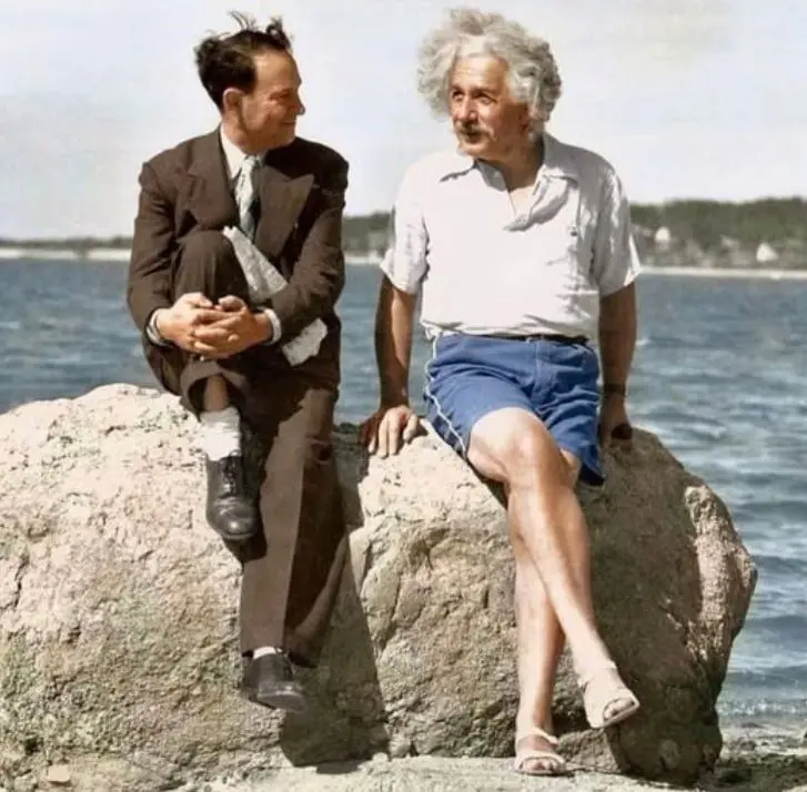 Two men sit on large rocks by the water. The man on the left, in a suit, looks at the man on the right, Albert Einstein, who…