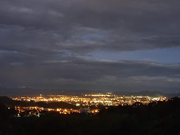 A wide-angle photograph of a sprawling cityscape at dusk or night, with countless warm yellow lights from buildings and…
