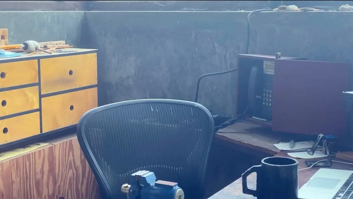 A dimly lit, cluttered office space with a black ergonomic chair centered in the frame. On the desk to the right sits a…