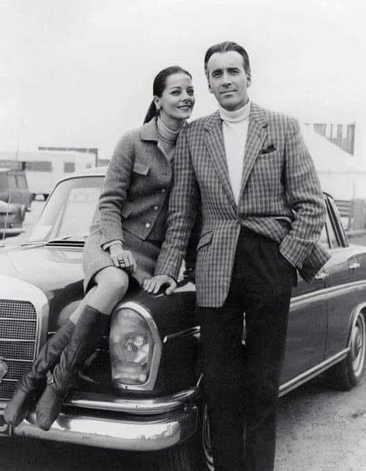 A black-and-white photo shows a man and woman posing beside a vintage Mercedes-Benz. The woman sits on the car's hood…