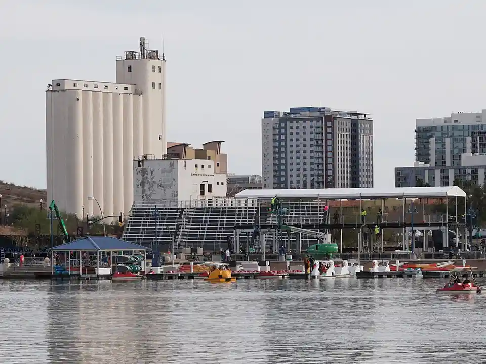 A wide shot of a waterfront with paddle boats and swan boats on the water, construction workers on scaffolding, and a large…