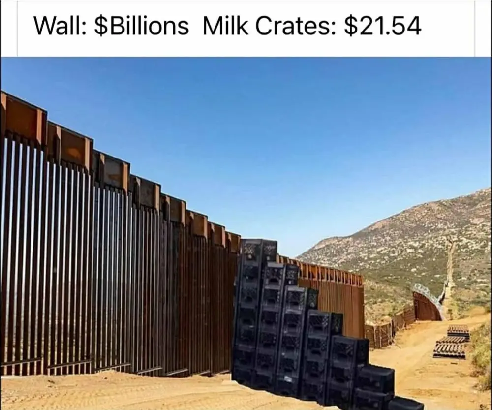 A long, tall metal border wall stretches across a desert landscape under a clear blue sky. Leaning against the wall is a…