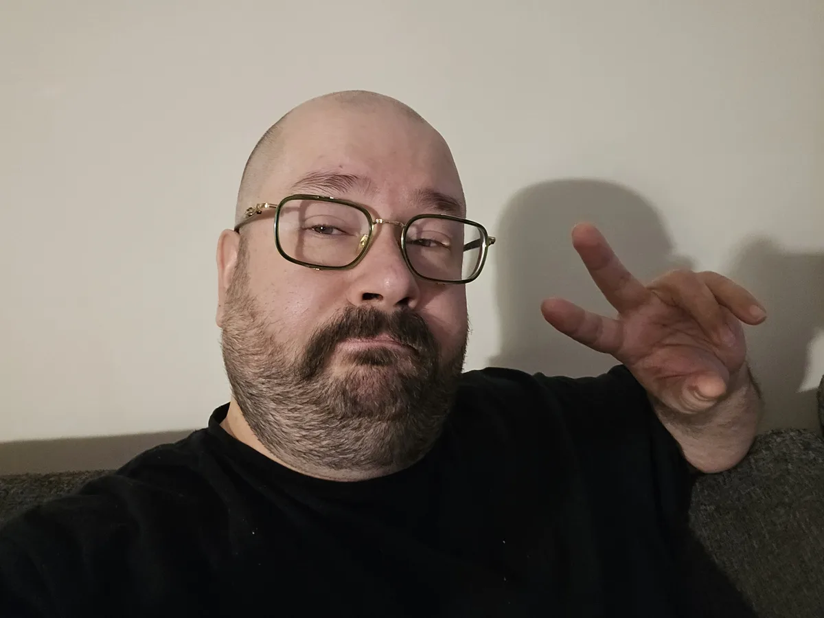 A bald man with a beard and gold-rimmed glasses sits on a couch, making a peace sign with his hand while looking at the…
