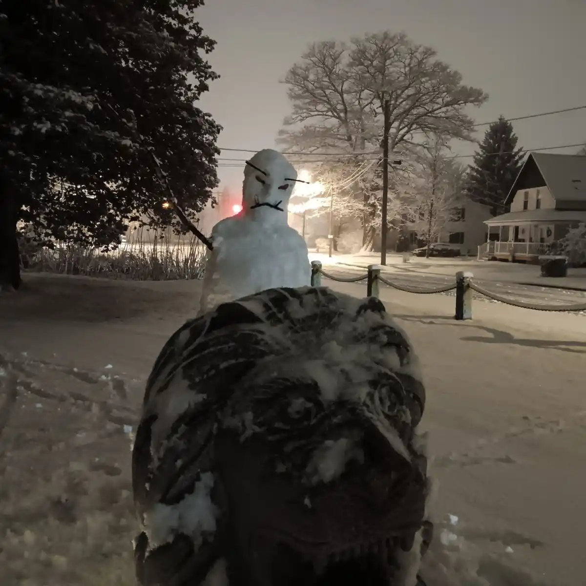 A snowman with an angry, menacing face made of sticks for eyes and mouth stands behind a snow-covered stone dinosaur head in…