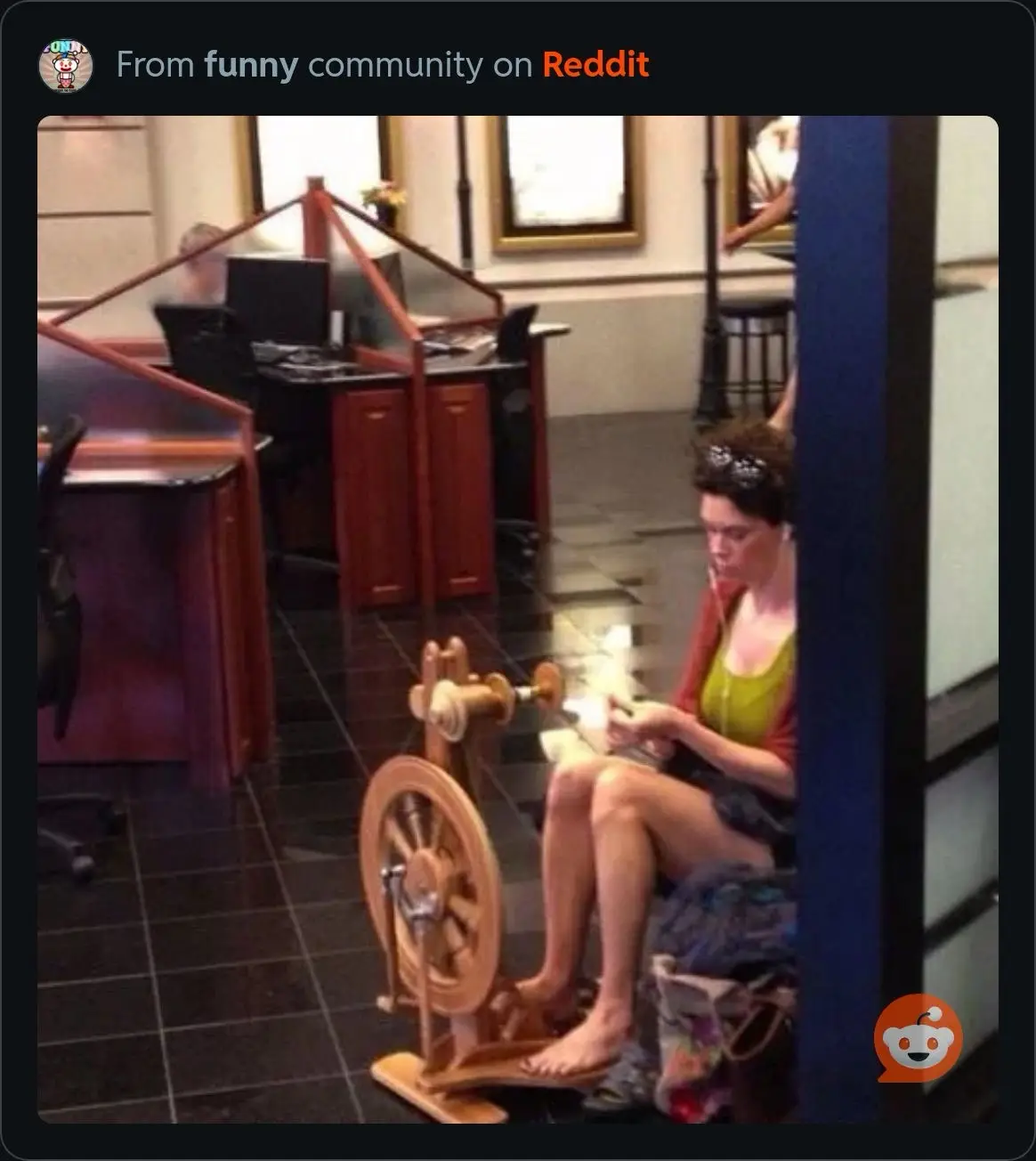 A woman sits on the floor in an office, barefoot, operating a wooden spinning wheel while looking down at her work. She…