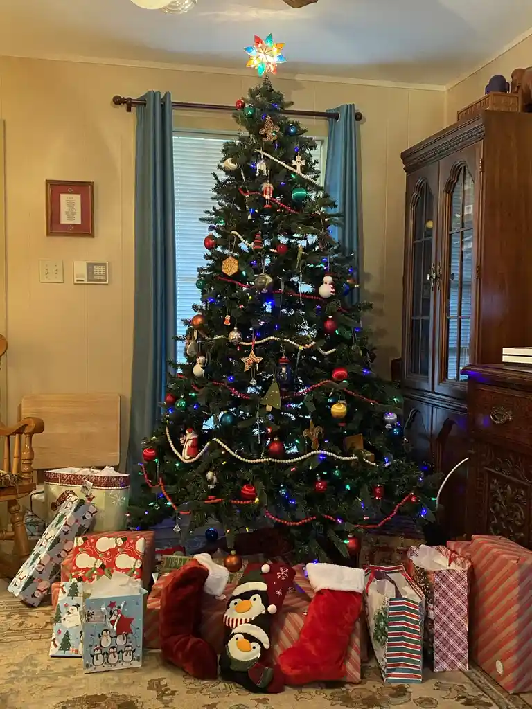A brightly lit, heavily decorated Christmas tree stands in a living room, surrounded by numerous wrapped gifts and red…