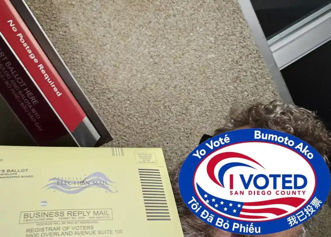 A close-up photo shows a yellow official election mail ballot envelope and a red ballot return envelope on a beige carpet. A…