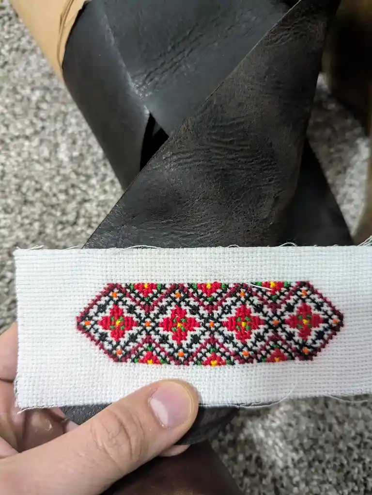 A hand holds a small white fabric swatch with a colorful, intricate cross-stitch pattern featuring red, black, green, and…