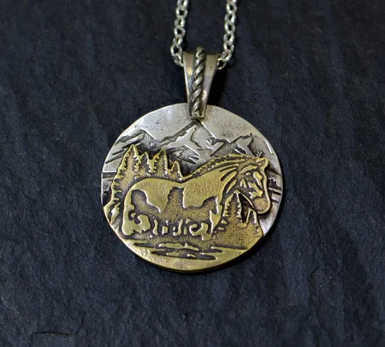 A close-up photo of a round, two-toned metal pendant necklace resting on a dark slate surface. The pendant features an…
