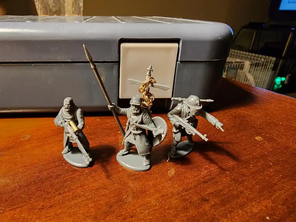 Three unpainted miniature soldier figures stand on a wooden table in front of a gray plastic case. The central figure holds…