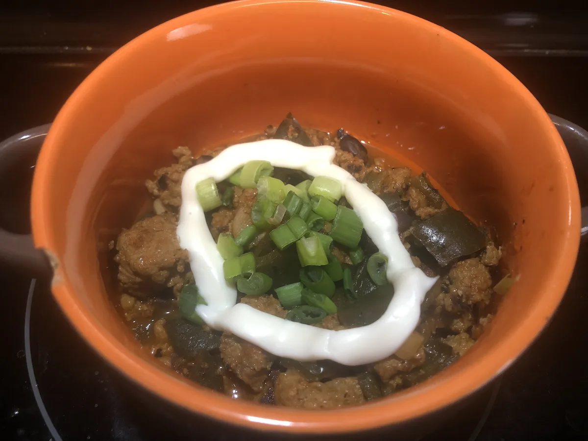 A close-up photo shows an orange ceramic bowl filled with a stew-like dish containing ground meat, dark green peppers, and…