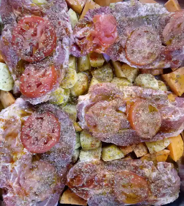A close-up photo shows raw meat pieces, likely pork chops, arranged over chopped sweet potatoes and other vegetables in a…