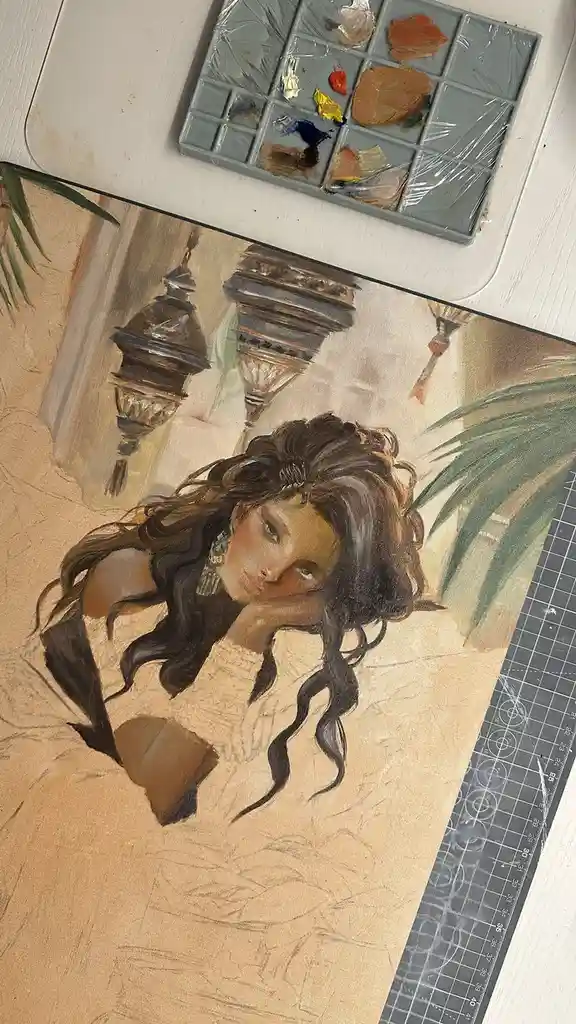 A partially painted artwork of a woman with long dark hair resting her head on her hand, set on a white surface next to a…