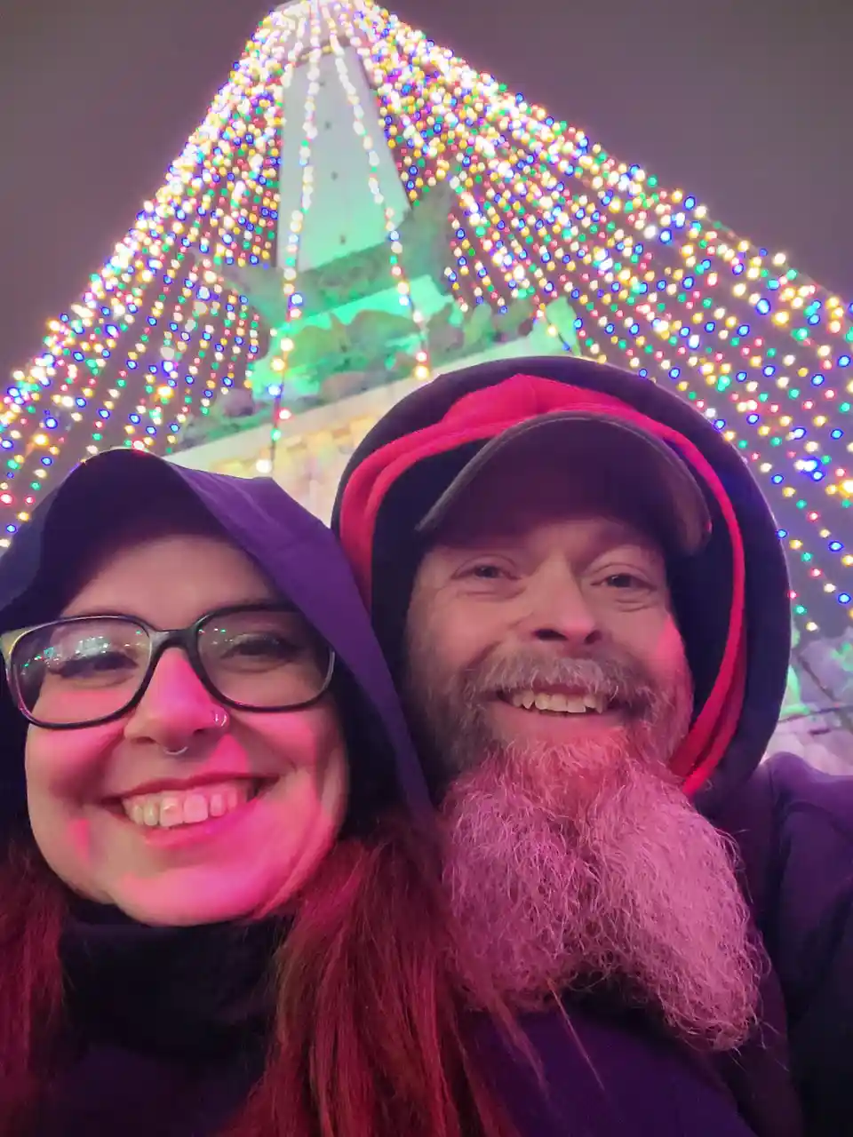 A selfie of a smiling man and woman in hoodies, taken at night in front of a brightly lit, colorful, triangular structure…