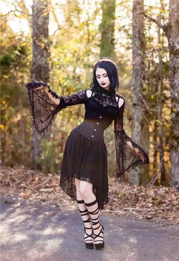 A woman with dark hair and gothic makeup stands in a forest path wearing a black lace dress with a corset, high heels, and…