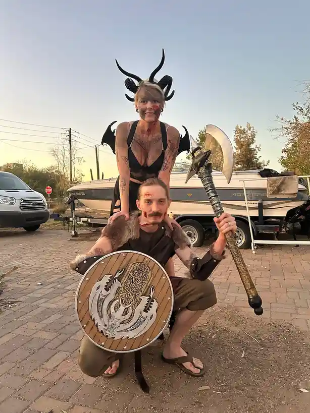 A man and woman dressed in fantasy warrior costumes with fake blood, horns, and wings pose outdoors near a boat and van. The…