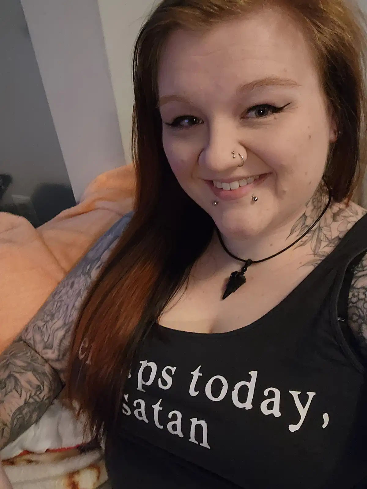 A smiling woman with long brown hair, multiple facial piercings, and tattoos on her neck and arms wears a black tank top…