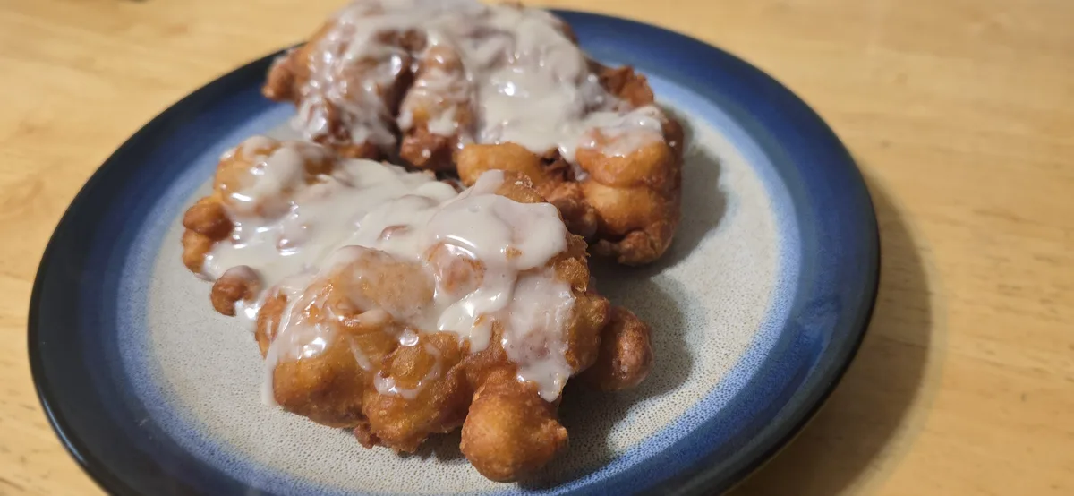 A close-up photo shows two golden-brown, fried, doughy pastries covered in white icing on a blue and white speckled plate…