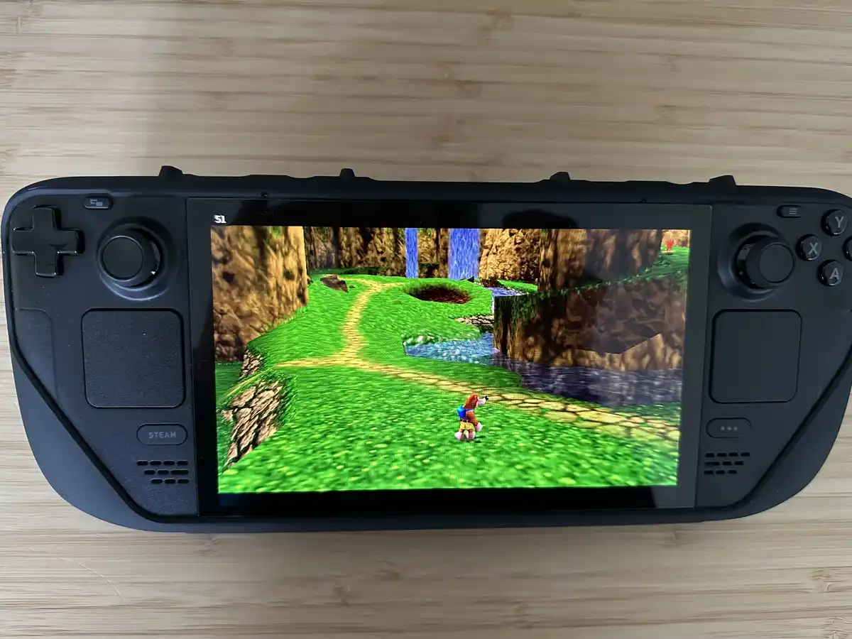A black Steam Deck handheld gaming console rests on a wooden surface, its screen displaying a 3D video game scene with a…