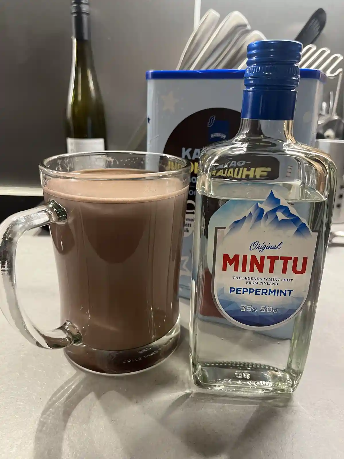 A glass mug of hot chocolate sits next to a bottle of Minttu peppermint schnapps on a kitchen counter. The bottle label…
