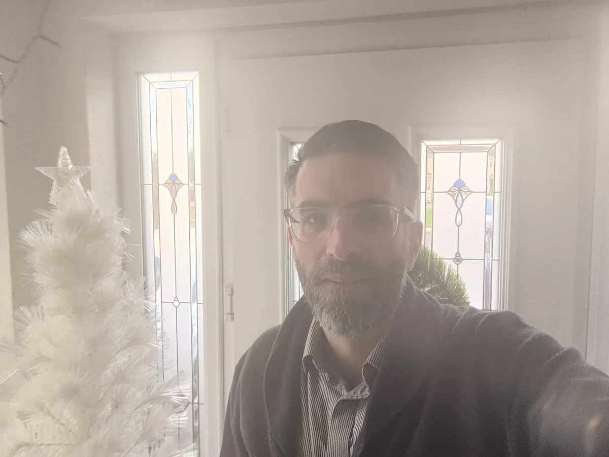 A man with glasses and a beard takes a selfie indoors, standing beside a white artificial Christmas tree topped with a star…