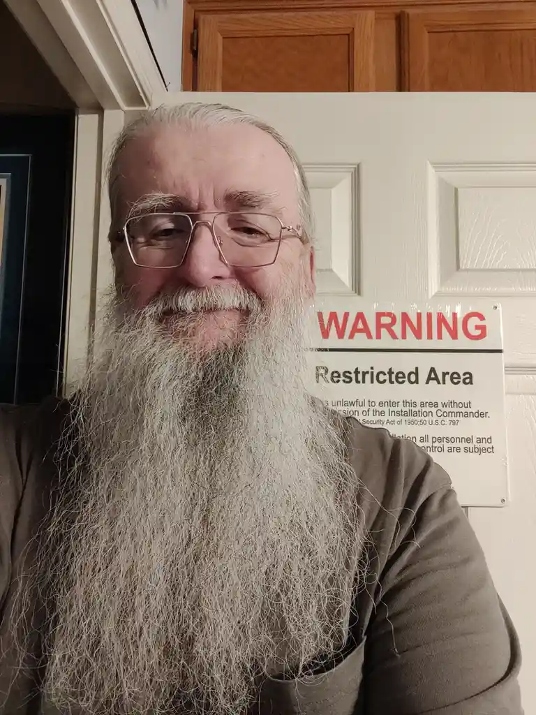 A man with a long white beard and glasses stands in front of a white door, smiling. A 'WARNING Restricted Area' sign is…