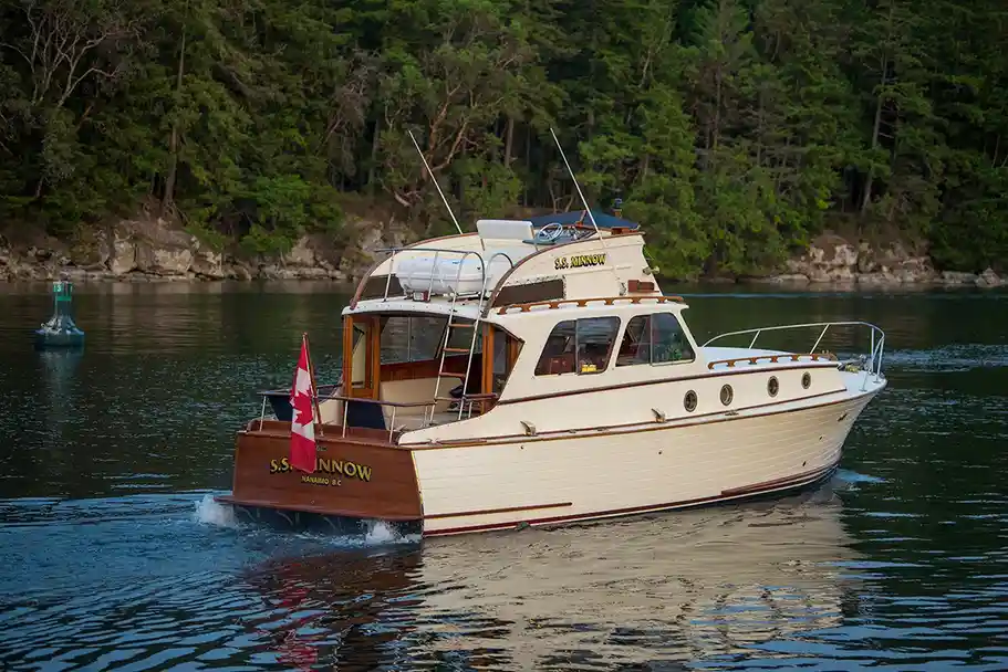 A cream-and-wood motor yacht named 'S.S. MINNOW' is shown cruising on calm water, with a Canadian flag flying from its stern…