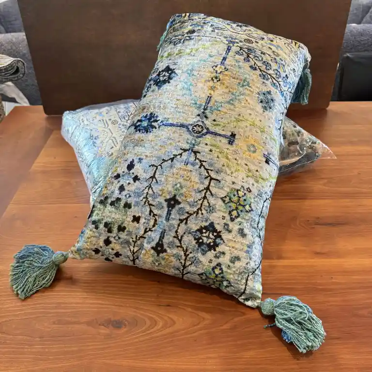 A decorative pillow with a blue, green, and beige floral and geometric pattern and tassels sits on a wooden table, partially…