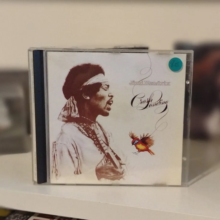 A CD case for Jimi Hendrix's album 'Crash Landing' sits on a shelf, featuring a sepia-toned image of Hendrix playing guitar…