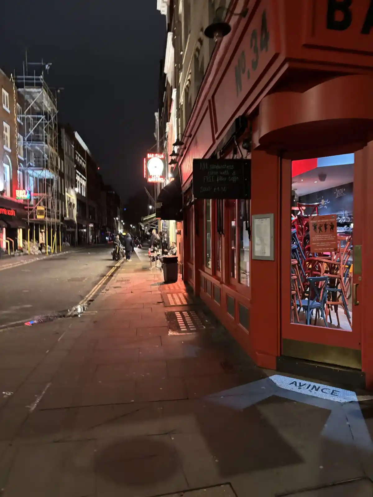 A nighttime street view of a red-painted bar or cafe named 'No. 34' with a chalkboard sign advertising 'All sandwiches come…