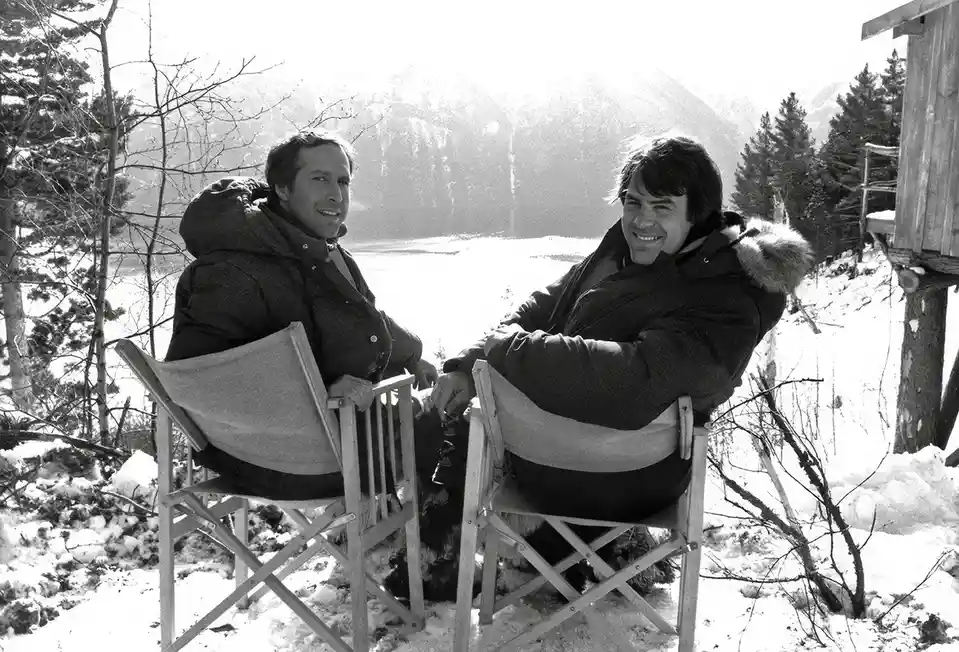 A black-and-white photo shows two men sitting in director's chairs outdoors in a snowy landscape with mountains and trees in…