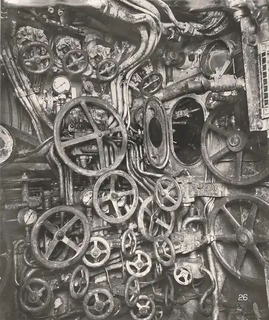 A black-and-white historical photograph shows a densely packed, complex interior of a ship's engine room, filled with…
