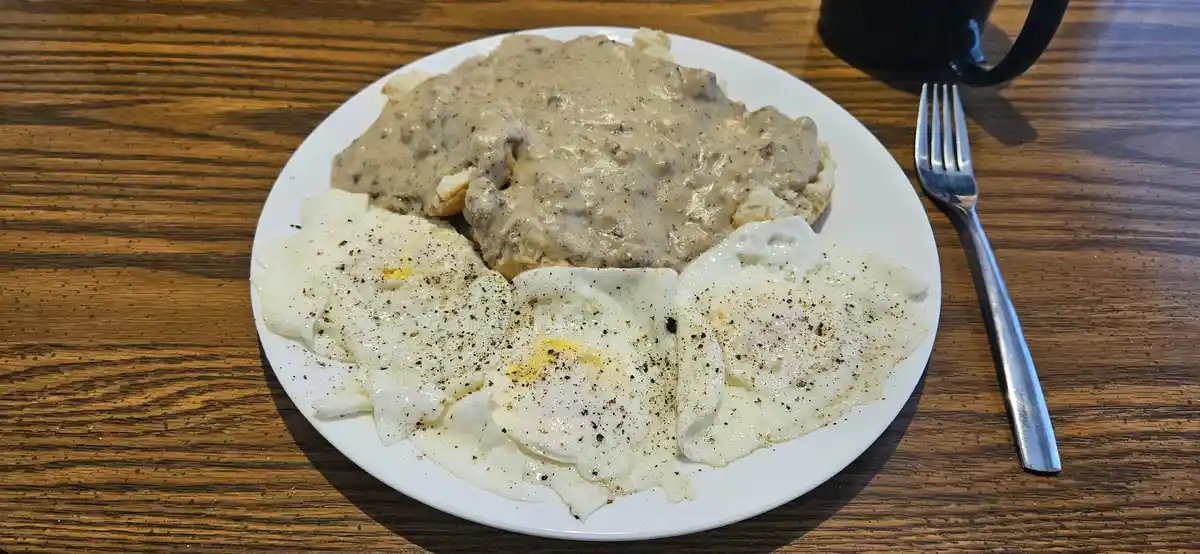 A plate of breakfast food on a wooden table, featuring three fried eggs seasoned with black pepper and a serving of biscuits…