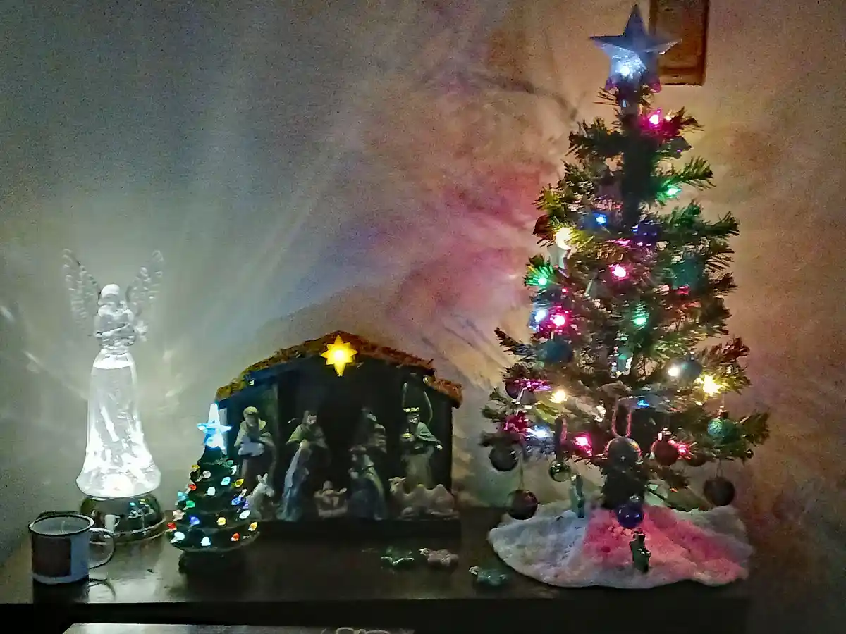 A decorated christmas tree with colorful lights and ornaments sits next to a nativity scene, which also contains a small…