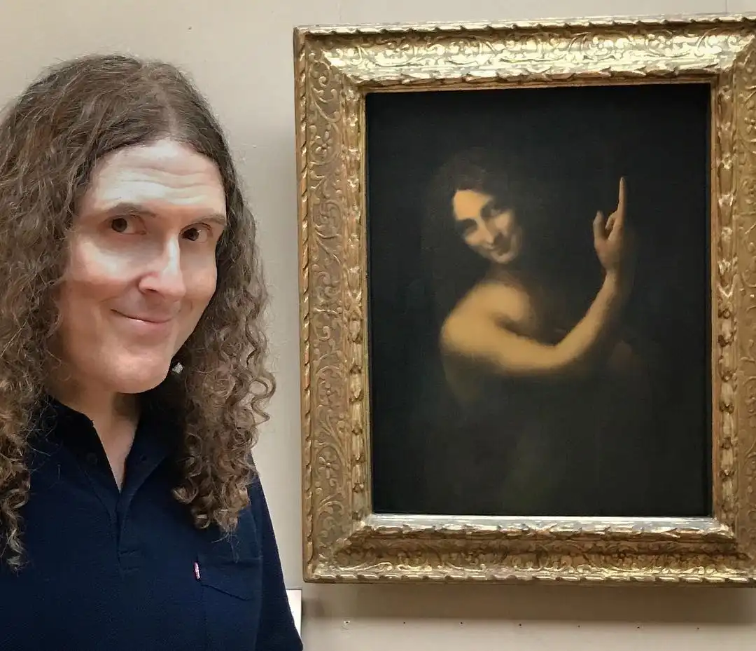 A man with long curly hair and a smug smile stands beside a framed painting of a woman with a similar smug expression, also…