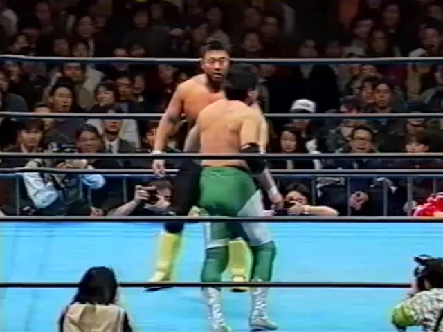 Two shirtless male wrestlers are in a ring, one in green trunks and boots is being held by the other in black trunks and…