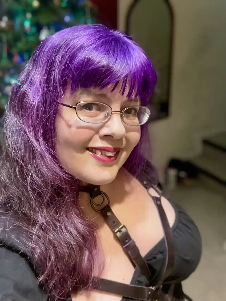 A woman with long purple hair and glasses smiles at the camera while wearing a black outfit with leather straps and a…