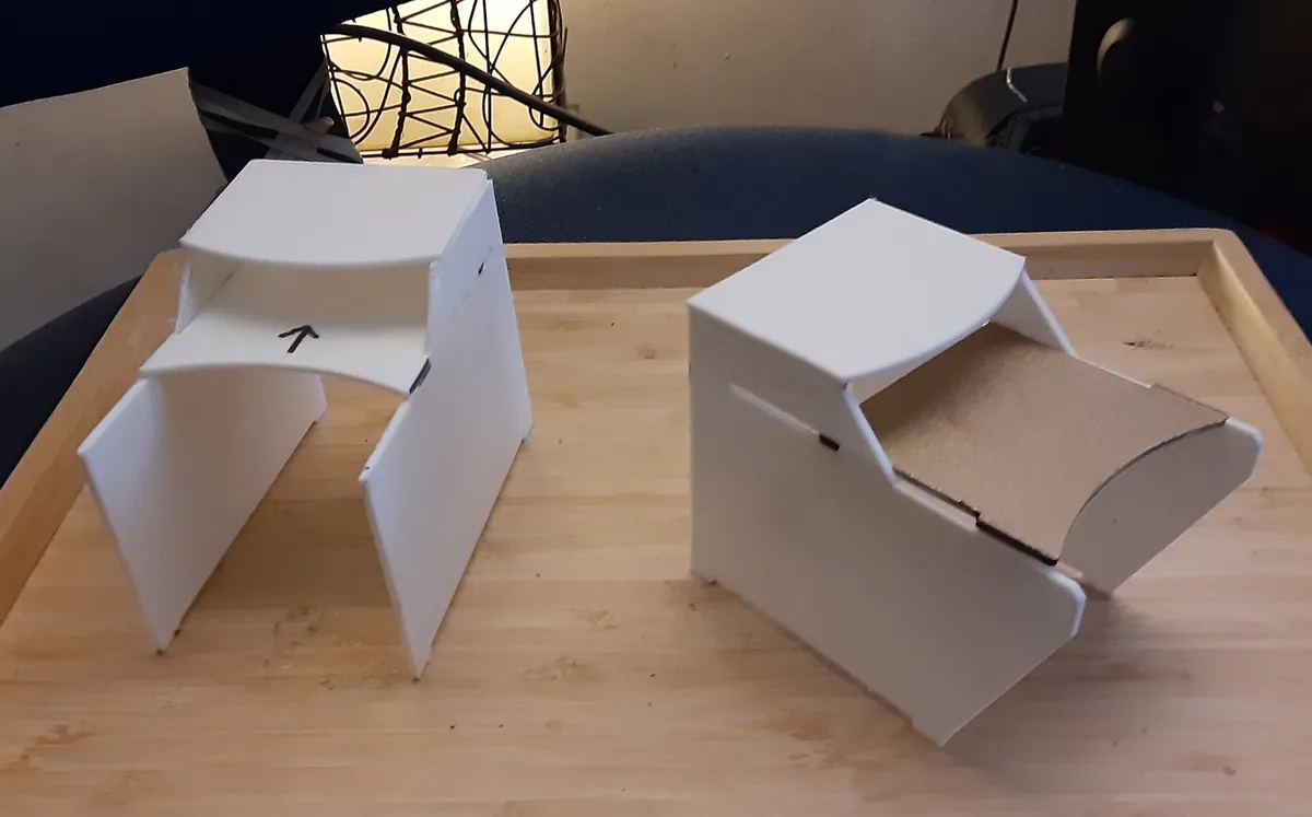 Two white cardboard box structures sit on a wooden tray, resembling chairs but clearly not functional ones. The left…