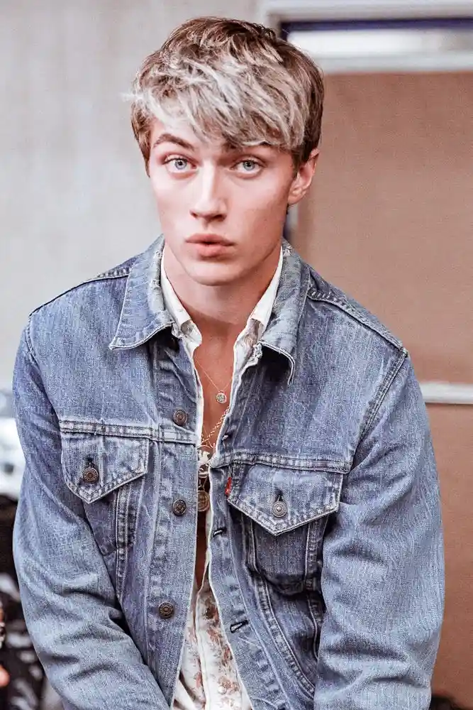 A young man with bleached blonde hair and blue eyes stares intently at the camera, wearing a denim jacket over a patterned…