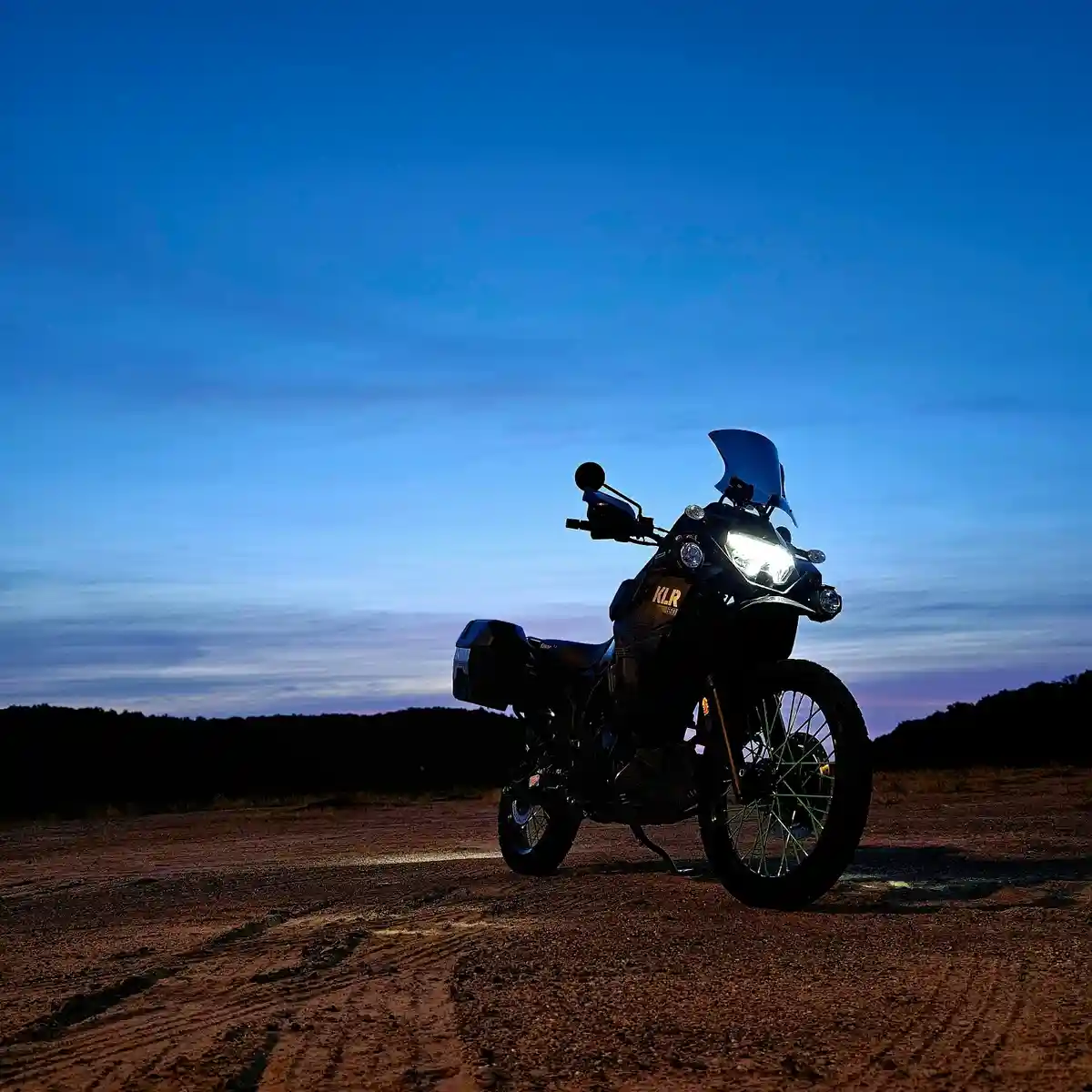 A black adventure motorcycle with its headlights on is parked on a dirt road at dusk, casting light on the ground. The sky…