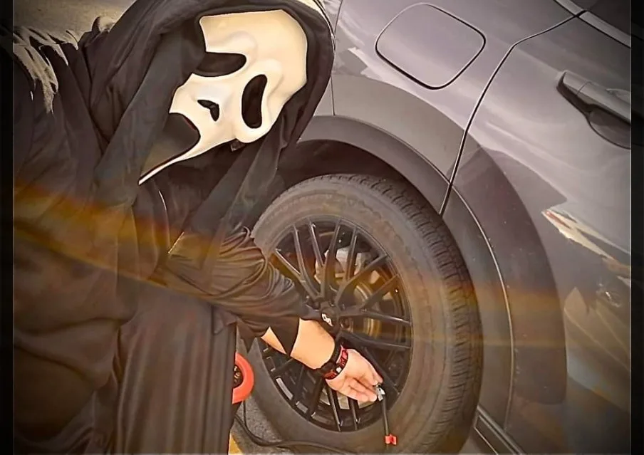 A person wearing a white Ghostface mask from the Scream movie franchise is shown leaning over the front wheel of a dark gray…