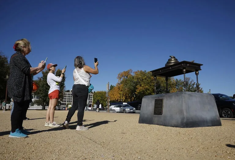 three women are standing in front of a public monument shaped like a giant metal toilet seat with a bird perched on top, all…